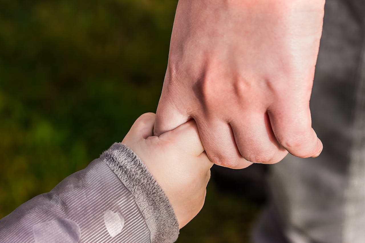about-us-01 A close-up of a child and parent holding hands in a park, symbolizing love and trust.