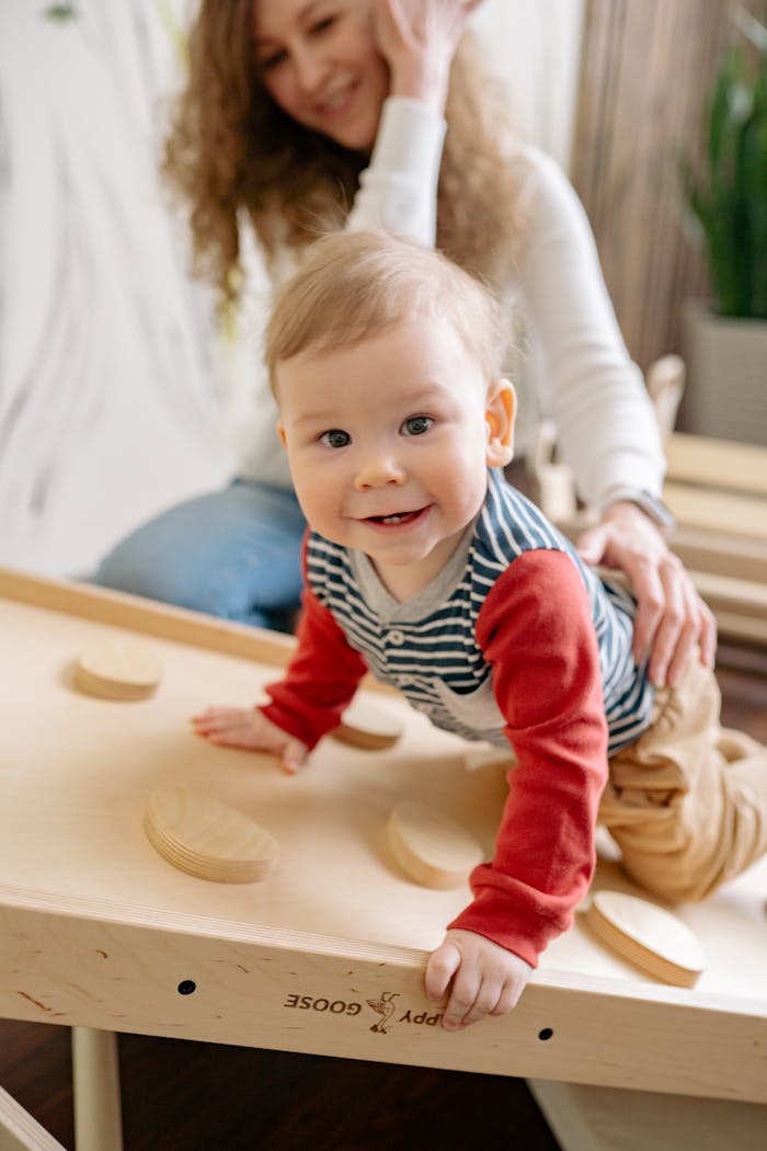Adorable baby crawling playfully on a wooden toy table with supportive mother nearby, indoors.